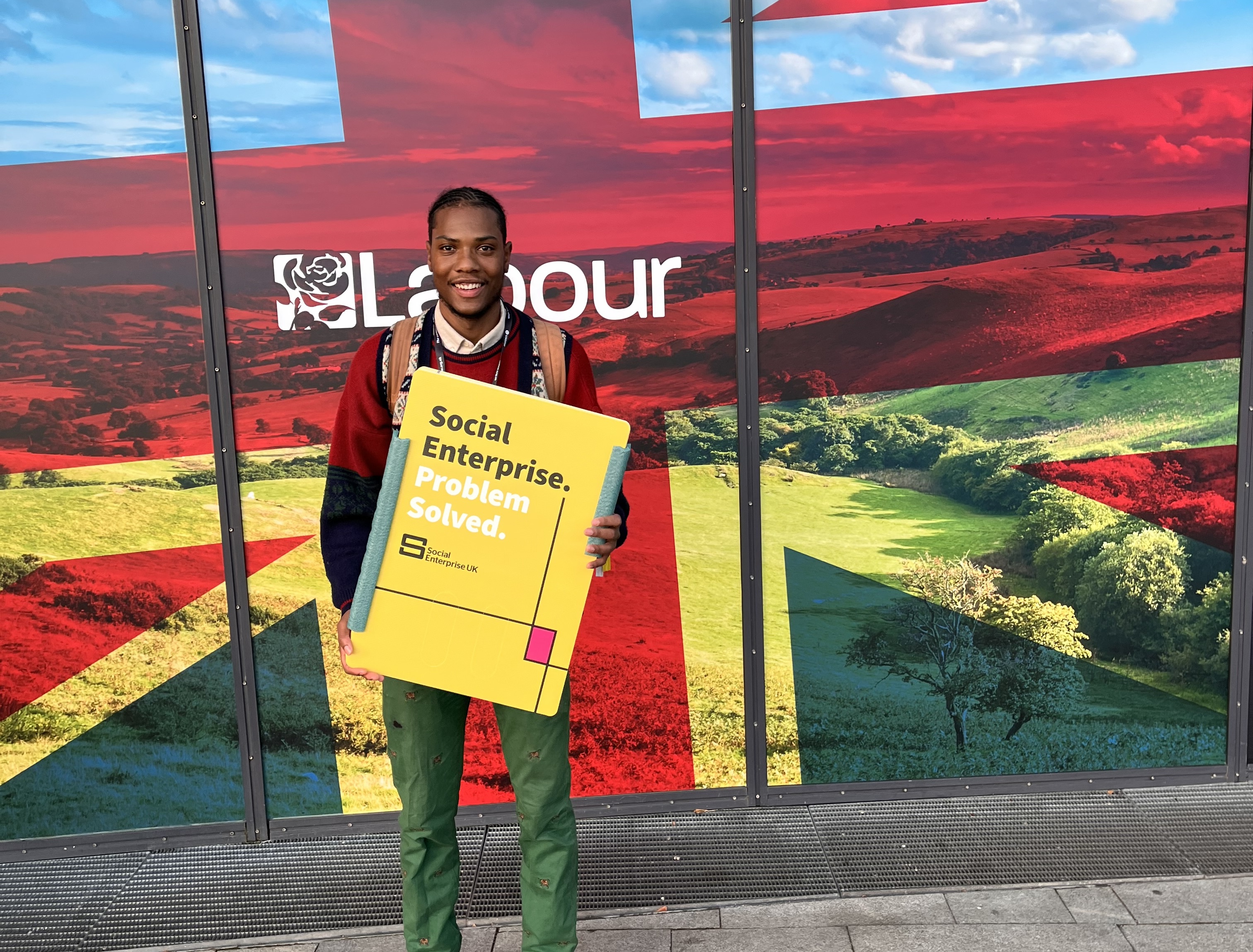 Jovan in front of a Labour Party logo holding a Social Enterprise UK sign