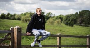 A young man sits on a wooden fence in a lush green field. He wears a black hoodie and grey joggers, with white trainers.